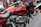 A close-up view of a red motorcycle with chrome details and branding visible on the fuel tank. The bike is positioned indoors, possibly in a showroom. Surrounding motorcycles are partially visible, adding depth to the scene.