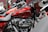 A close-up view of a red motorcycle with chrome details and branding visible on the fuel tank. The bike is positioned indoors, possibly in a showroom. Surrounding motorcycles are partially visible, adding depth to the scene.