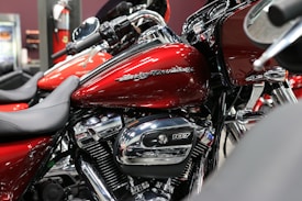 A close-up view of a red motorcycle with chrome details and branding visible on the fuel tank. The bike is positioned indoors, possibly in a showroom. Surrounding motorcycles are partially visible, adding depth to the scene.