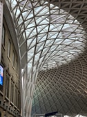 A spacious, modern architectural interior showcasing a large, curving roof structure made of interwoven steel beams set against a glass ceiling. The left side features a brick facade with arched windows and a digital screen displaying advertisements. The design blends historic and contemporary styles.