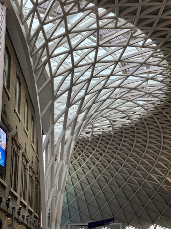 A spacious, modern architectural interior showcasing a large, curving roof structure made of interwoven steel beams set against a glass ceiling. The left side features a brick facade with arched windows and a digital screen displaying advertisements. The design blends historic and contemporary styles.