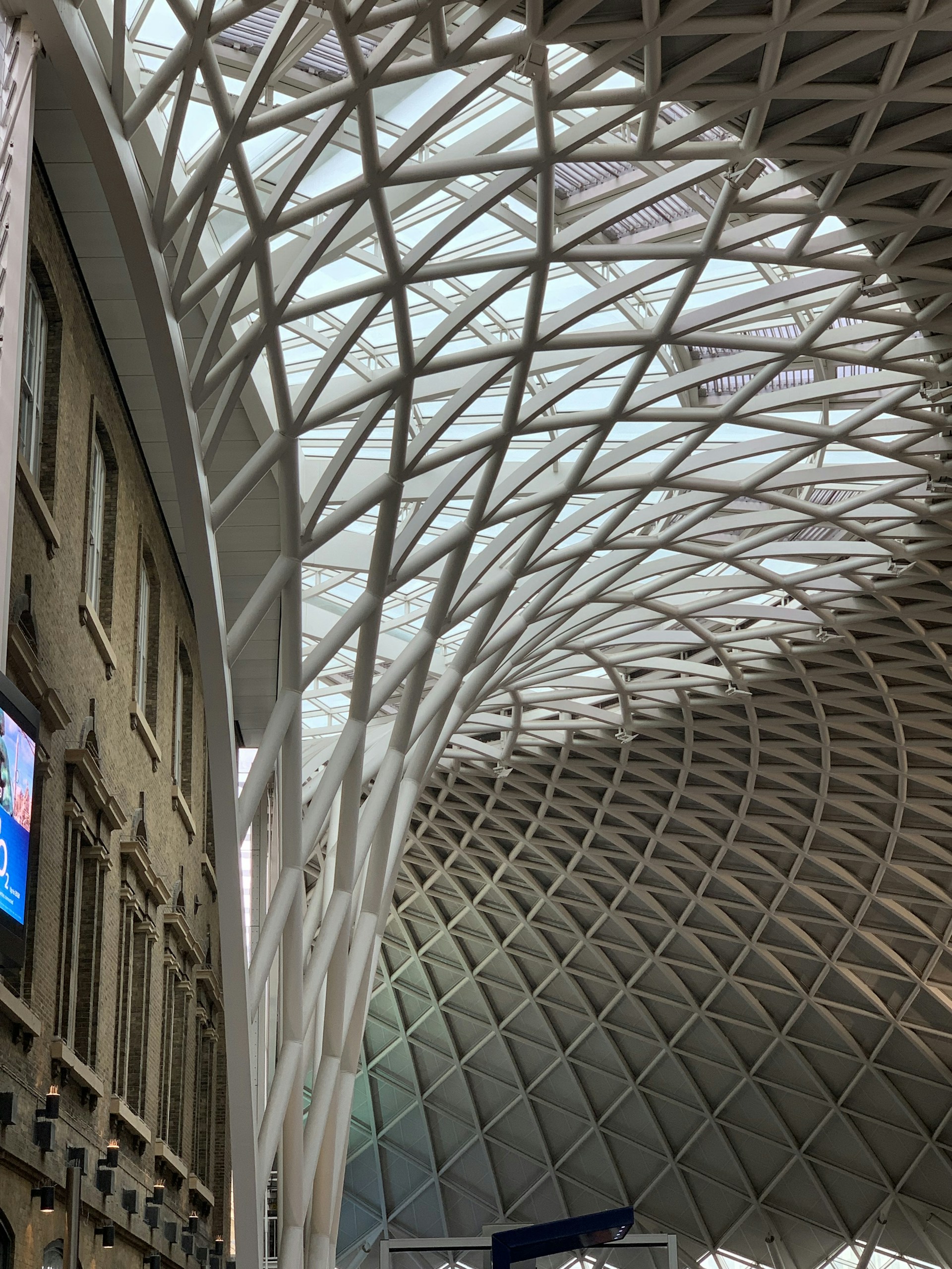 A spacious, modern architectural interior showcasing a large, curving roof structure made of interwoven steel beams set against a glass ceiling. The left side features a brick facade with arched windows and a digital screen displaying advertisements. The design blends historic and contemporary styles.
