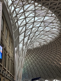 A spacious, modern architectural interior showcasing a large, curving roof structure made of interwoven steel beams set against a glass ceiling. The left side features a brick facade with arched windows and a digital screen displaying advertisements. The design blends historic and contemporary styles.