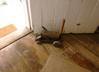 A small, rustic wooden toy vehicle with wheels rests on an aged wooden floor near a white wooden door. The toy appears handcrafted, with a simple design and a rod protruding from its base.