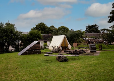 A rustic outdoor campsite featuring a white canvas tent is set up on a grassy field surrounded by trees. A simple wooden structure is nearby, along with a wooden barrel and benches. A fire pit with logs and ashes sits in front of the tent, suggesting recent use. In the background, there are traditional wooden buildings that add to the historical or rural feel.