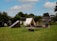 A rustic outdoor campsite featuring a white canvas tent is set up on a grassy field surrounded by trees. A simple wooden structure is nearby, along with a wooden barrel and benches. A fire pit with logs and ashes sits in front of the tent, suggesting recent use. In the background, there are traditional wooden buildings that add to the historical or rural feel.