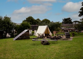 A rustic outdoor campsite featuring a white canvas tent is set up on a grassy field surrounded by trees. A simple wooden structure is nearby, along with a wooden barrel and benches. A fire pit with logs and ashes sits in front of the tent, suggesting recent use. In the background, there are traditional wooden buildings that add to the historical or rural feel.