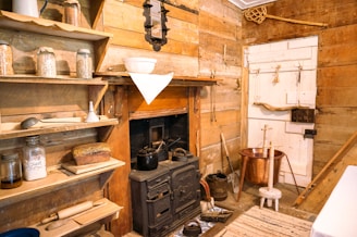 A rustic kitchen with wooden walls and shelves filled with jars of grains and a container labeled 'Salt'. A black stove is placed against the wall, accompanied by various kitchen utensils. A loaf of bread rests on a wooden shelf beside a rolling pin and jars. The interior features warm wooden tones with a copper pot near the door, adding to the cozy, vintage ambiance.