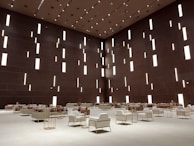 A spacious modern interior features a high ceiling with numerous vertical rectangular light openings. The walls have a dark brown tone, creating a sophisticated and minimalist ambiance. The room is furnished with organized rows of plush armchairs and sofas, predominantly in white and soft taupe colors, complemented by metallic side tables. The arrangement reflects a balance between luxury and minimalism.