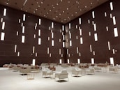 A spacious modern interior features a high ceiling with numerous vertical rectangular light openings. The walls have a dark brown tone, creating a sophisticated and minimalist ambiance. The room is furnished with organized rows of plush armchairs and sofas, predominantly in white and soft taupe colors, complemented by metallic side tables. The arrangement reflects a balance between luxury and minimalism.