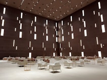 A spacious modern interior features a high ceiling with numerous vertical rectangular light openings. The walls have a dark brown tone, creating a sophisticated and minimalist ambiance. The room is furnished with organized rows of plush armchairs and sofas, predominantly in white and soft taupe colors, complemented by metallic side tables. The arrangement reflects a balance between luxury and minimalism.