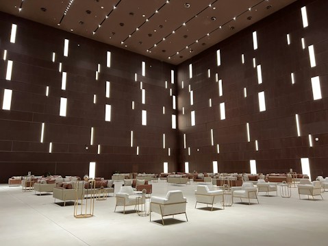 A spacious modern interior features a high ceiling with numerous vertical rectangular light openings. The walls have a dark brown tone, creating a sophisticated and minimalist ambiance. The room is furnished with organized rows of plush armchairs and sofas, predominantly in white and soft taupe colors, complemented by metallic side tables. The arrangement reflects a balance between luxury and minimalism.