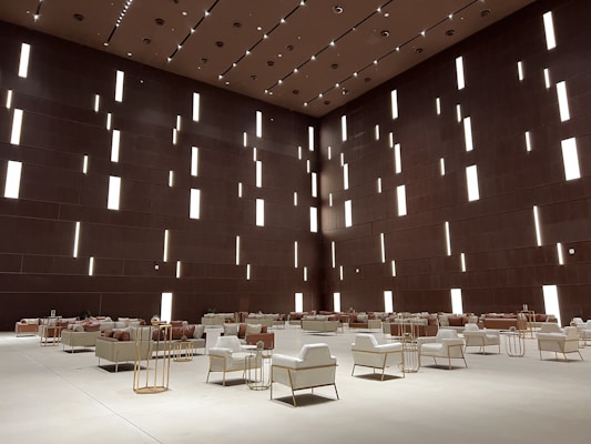 A spacious modern interior features a high ceiling with numerous vertical rectangular light openings. The walls have a dark brown tone, creating a sophisticated and minimalist ambiance. The room is furnished with organized rows of plush armchairs and sofas, predominantly in white and soft taupe colors, complemented by metallic side tables. The arrangement reflects a balance between luxury and minimalism.