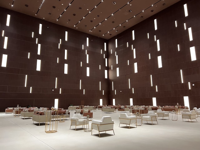 A spacious modern interior features a high ceiling with numerous vertical rectangular light openings. The walls have a dark brown tone, creating a sophisticated and minimalist ambiance. The room is furnished with organized rows of plush armchairs and sofas, predominantly in white and soft taupe colors, complemented by metallic side tables. The arrangement reflects a balance between luxury and minimalism.