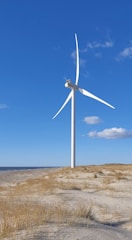 A large white wind turbine stands on a sandy coastal landscape with sparse grass and a clear blue sky. The horizon shows a vast ocean and a few distant clouds.