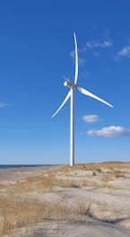 A large white wind turbine stands on a sandy coastal landscape with sparse grass and a clear blue sky. The horizon shows a vast ocean and a few distant clouds.