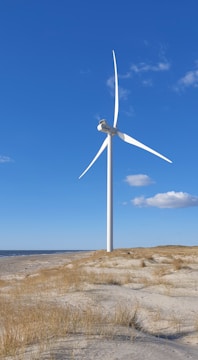 A large white wind turbine stands on a sandy coastal landscape with sparse grass and a clear blue sky. The horizon shows a vast ocean and a few distant clouds.