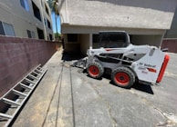 A small white Bobcat skid-steer loader is parked on a paved surface beside a building. The loader has red wheels and is positioned near a brick wall and a ladder lying on the ground. There are two beige multi-story buildings with windows nearby, and sunlight casts shadows on the ground.
