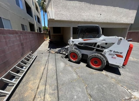 A small white Bobcat skid-steer loader is parked on a paved surface beside a building. The loader has red wheels and is positioned near a brick wall and a ladder lying on the ground. There are two beige multi-story buildings with windows nearby, and sunlight casts shadows on the ground.