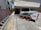 A small white Bobcat skid-steer loader is parked on a paved surface beside a building. The loader has red wheels and is positioned near a brick wall and a ladder lying on the ground. There are two beige multi-story buildings with windows nearby, and sunlight casts shadows on the ground.