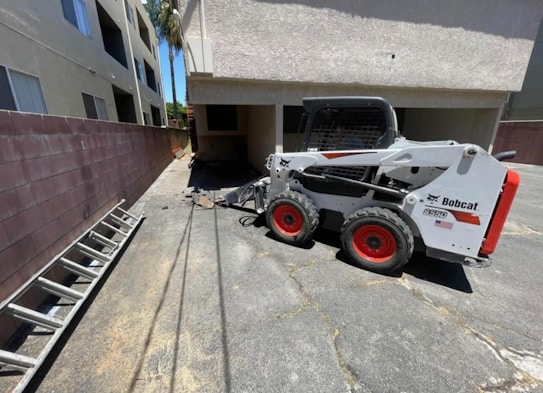 A small white Bobcat skid-steer loader is parked on a paved surface beside a building. The loader has red wheels and is positioned near a brick wall and a ladder lying on the ground. There are two beige multi-story buildings with windows nearby, and sunlight casts shadows on the ground.