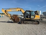 A large yellow construction excavator with a prominent digging arm and bucket is parked on a gravel surface. The machine has rubber tracks and is part of a construction site with various industrial items and buildings visible in the background.