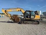 A large yellow construction excavator with a prominent digging arm and bucket is parked on a gravel surface. The machine has rubber tracks and is part of a construction site with various industrial items and buildings visible in the background.