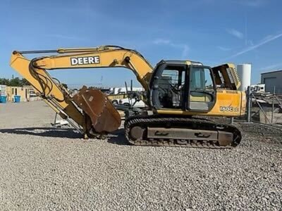A large yellow construction excavator with a prominent digging arm and bucket is parked on a gravel surface. The machine has rubber tracks and is part of a construction site with various industrial items and buildings visible in the background.