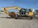 A large yellow construction excavator with a prominent digging arm and bucket is parked on a gravel surface. The machine has rubber tracks and is part of a construction site with various industrial items and buildings visible in the background.
