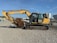 A large yellow construction excavator with a prominent digging arm and bucket is parked on a gravel surface. The machine has rubber tracks and is part of a construction site with various industrial items and buildings visible in the background.