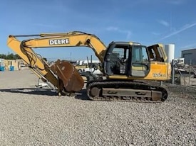 A large yellow construction excavator with a prominent digging arm and bucket is parked on a gravel surface. The machine has rubber tracks and is part of a construction site with various industrial items and buildings visible in the background.