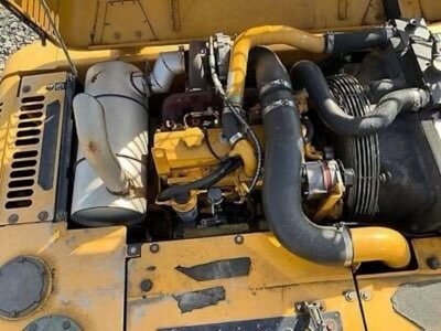 A detailed view of a yellow industrial engine with various components, including hoses, a fan belt, and metallic parts. There is a prominent use of black and yellow materials, and the engine is set against a sunny background which casts shadows on its surface.