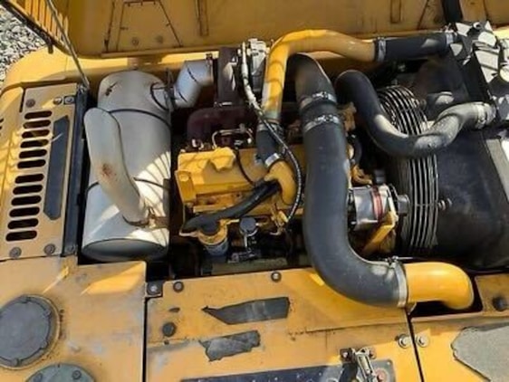 A detailed view of a yellow industrial engine with various components, including hoses, a fan belt, and metallic parts. There is a prominent use of black and yellow materials, and the engine is set against a sunny background which casts shadows on its surface.