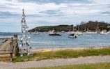 A coastal scene featuring a bay with several boats anchored on the water. The shore is lined with a grassy area and a paved path. In the foreground, a decorative structure made from seashells and wooden pieces is positioned near a wooden fence. The backdrop includes a distant hilly landscape covered with trees.
