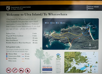A signboard presents information about Ulva Island/Te Wharawhara, a pest-free open island sanctuary in New Zealand. It includes a map showing various trails and points of interest, such as Sydney Cove, Flagstaff Point, and Boulder Beach. The board lists walking times and types of self-guided walks (history, conservation, and nature). Contact information for the Rakiura National Park Visitor Centre is provided for pest sightings and general inquiries. Symbols indicating prohibited actions, such as no dogs, are displayed at the bottom.