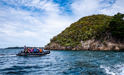 A group of people wearing life jackets is seated on an inflatable boat traveling on a body of water. In the background, a lush green island with rocky outcrops is visible under a partly cloudy sky.