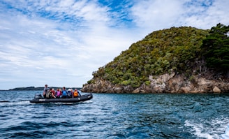 A group of people wearing life jackets is seated on an inflatable boat traveling on a body of water. In the background, a lush green island with rocky outcrops is visible under a partly cloudy sky.
