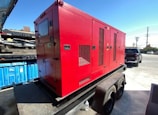 A large red generator is mounted on a trailer, parked in an industrial area with a blue dumpster nearby. In the background, a black pickup truck and a utility pole are visible under a clear blue sky. The ground is paved with concrete.