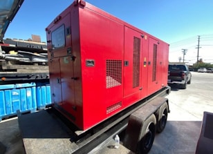 A large red generator is mounted on a trailer, parked in an industrial area with a blue dumpster nearby. In the background, a black pickup truck and a utility pole are visible under a clear blue sky. The ground is paved with concrete.