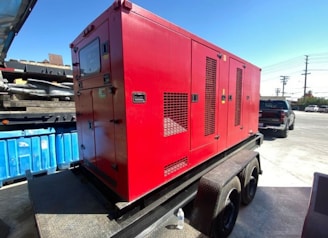 A large red generator is mounted on a trailer, parked in an industrial area with a blue dumpster nearby. In the background, a black pickup truck and a utility pole are visible under a clear blue sky. The ground is paved with concrete.