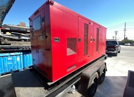 A large red generator is mounted on a trailer, parked in an industrial area with a blue dumpster nearby. In the background, a black pickup truck and a utility pole are visible under a clear blue sky. The ground is paved with concrete.