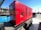 A large red generator is mounted on a trailer, parked in an industrial area with a blue dumpster nearby. In the background, a black pickup truck and a utility pole are visible under a clear blue sky. The ground is paved with concrete.