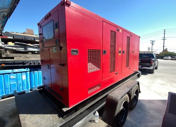 A large red generator is mounted on a trailer, parked in an industrial area with a blue dumpster nearby. In the background, a black pickup truck and a utility pole are visible under a clear blue sky. The ground is paved with concrete.