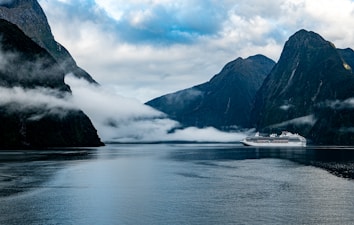 A large cruise ship travels through a calm fjord surrounded by towering dark mountains partially shrouded in mist and low clouds. The serene water reflects the dramatic landscape, creating an atmosphere of tranquility.