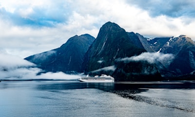 A large cruise ship sails through calm waters, flanked by towering, rugged mountains partially shrouded in mist and clouds. The scene captures a dramatic contrast between the man-made ship and the natural grandeur of the landscape, with the peaks stretching high into a cloudy sky.