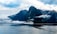 A large cruise ship sails through calm waters, flanked by towering, rugged mountains partially shrouded in mist and clouds. The scene captures a dramatic contrast between the man-made ship and the natural grandeur of the landscape, with the peaks stretching high into a cloudy sky.