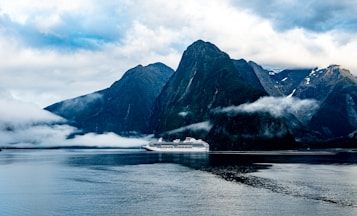 A large cruise ship sails through calm waters, flanked by towering, rugged mountains partially shrouded in mist and clouds. The scene captures a dramatic contrast between the man-made ship and the natural grandeur of the landscape, with the peaks stretching high into a cloudy sky.