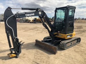 A compact excavator with a black and yellow body is parked on a dirt lot. The machinery has a large arm extending from its body, equipped with a digging bucket. The cabin is enclosed with large windows, and the tracks are visible beneath. In the background, there are other industrial vehicles and equipment visible, along with some structures and a cloudy sky.