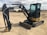 A compact excavator with a black and yellow body is parked on a dirt lot. The machinery has a large arm extending from its body, equipped with a digging bucket. The cabin is enclosed with large windows, and the tracks are visible beneath. In the background, there are other industrial vehicles and equipment visible, along with some structures and a cloudy sky.