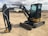 A compact excavator with a black and yellow body is parked on a dirt lot. The machinery has a large arm extending from its body, equipped with a digging bucket. The cabin is enclosed with large windows, and the tracks are visible beneath. In the background, there are other industrial vehicles and equipment visible, along with some structures and a cloudy sky.